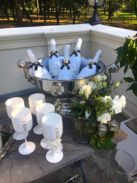 Elegancka dekoracja bufetu na tarasie z szampanem Moët & Chandon w lodzie, kieliszkami 'Moët', wazonem z białymi i zielonymi kwiatami oraz parkiem w tle