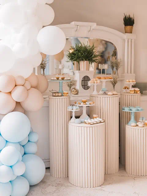 Elegancka dekoracja bufetu z białymi podstawkami, tacami z pastelowymi ciastkami i babeczkami, lustrem, doniczką z rośliną i balonami tworzącymi łuk