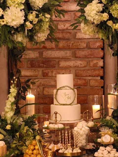 Bufet ślubny z elegancką dekoracją, biały tort Love w centrum, białe hortensje, zieleń, świece, makaroniki i inne słodycze, na tle ceglanej ściany
