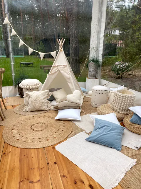 Wnętrze namiotu z dekoracją na komunię, tipi, poduszki w odcieniach bieli i beżu, plecione kosze, maty, girlanda z chorągiewek, posąg indianina w tle