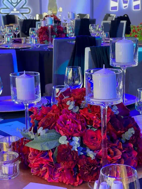Elegancka dekoracja stołu na imprezie firmowej w Warszawie z czerwonymi kwiatami, świecznikami, kieliszkami i talerzami, w tle sala z niebieskim oświetleniem