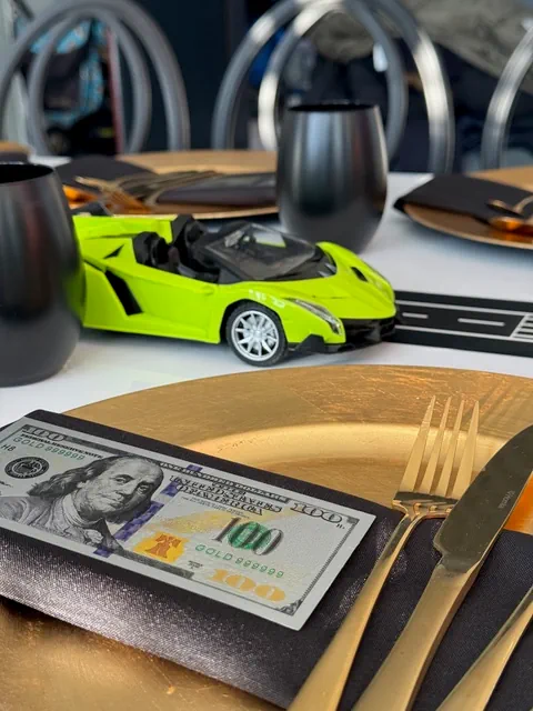 Elegancka dekoracja stołu na urodziny w Warszawie z złotymi elementami, czarnymi akcentami, modelem samochodu, banknotem 100 dolarów i stylowymi krzesłami