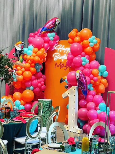 Kolorowa dekoracja urodzinowa w Warszawie: balony w odcieniach różu, pomarańczu i turkusu, papugi, napis Happy Birthday Maja, cyfra 1, kwiaty i eleganckie stoły