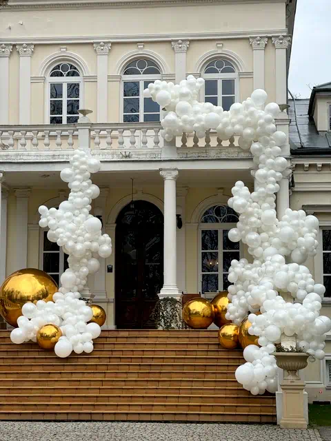 Dekoracja balonowa w kolorach białym i złotym, wijąca się wokół schodów i kolumn przed klasycznym budynkiem z łukowatymi oknami i balustradą