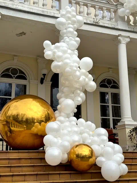 Elegancka dekoracja balonowa w kolorach bieli i złota przed wejściem do budynku z klasyczną architekturą, dodająca uroku i świątecznej atmosfery