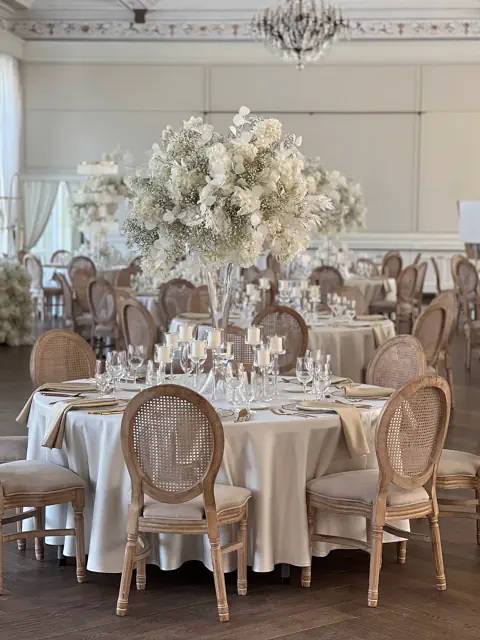 Sala balowa w Pałacu Mała Wieś z okrągłymi stołami, białymi obrusami i wysoką, białą dekoracją kwiatową. Świece, kieliszki, talerze i krzesła z plecionymi oparciami. Żyrandol i jasne ściany tworzą wyrafinowaną atmosferę przyjęcia