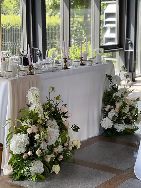 Elegancka dekoracja stołu w Pałacu Mała Wieś z białymi i różowymi kwiatami, szklanymi naczyniami i świecami, z dużymi oknami i widokiem na zieleń