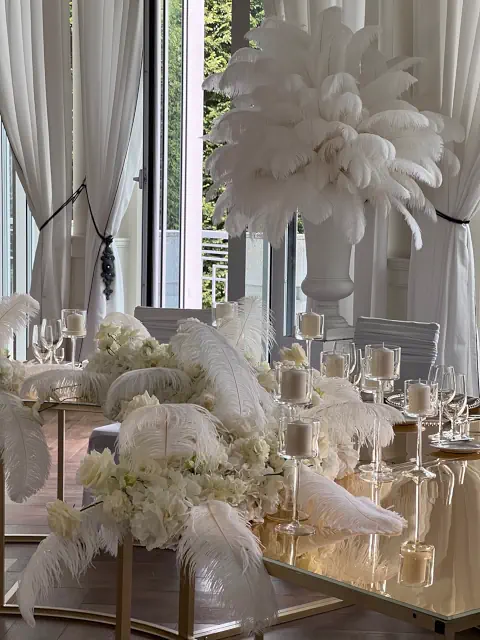 Elegancka dekoracja stołu w Pałacu Mała Wieś z białymi piórami, kwiatami, świecami, otoczona białymi zasłonami i dużymi oknami, z wysoką wazą pełną piór w tle