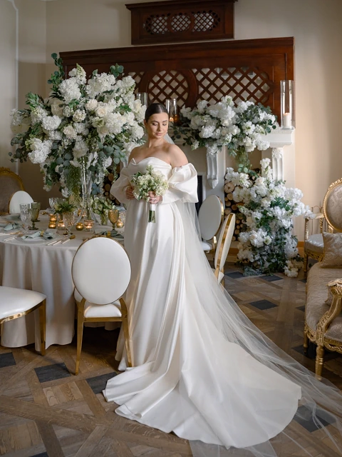 Panna młoda w białej sukni z welonem i bukietem w eleganckim wnętrzu z dekoracjami weselnymi w Warszawie. W tle kwiaty, stół, krzesła i drewniana ścianka