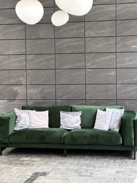 Ciemnozielona sofa welurowa na tle szarej, marmurowej ściany z panelami. Eleganckie poduszki i kuliste lampy jako dekoracja chillout w Warszawie