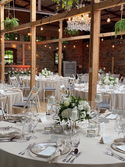 Elegancka sala weselna Cicha 23 Marki z drewnianą konstrukcją, ceglanymi ścianami i żyrandolem. Stoły przykryte białymi obrusami, ozdobione kwiatami i świecami, otoczono krzesłami z szarymi poduszkami. W tle widoczne rośliny i lampki, tworząc ciepłą atmosferę