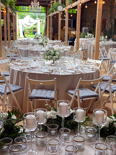 Elegancka sala weselna Cicha 23 Marki z okrągłymi stołami pokrytymi białymi obrusami, udekorowanymi kwiatami i świecami. Krzesła z szarymi poduszkami, żyrandole i zielone rośliny dodają uroku. W tle widoczne otwarte drzwi i dekoracja z tiulu