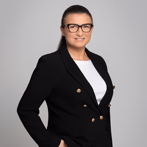 Marlena Kraska w czarnej marynarce, białej bluzce, z włosami w kucyku, okularami, uśmiechem, profesjonalnym wyglądem, ręką w kieszeni, z delikatną dekoracją na szyi
